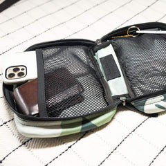 The GR1 Field Pocket x Gear Patrol from GORUCK, in Variegated Elephant Ear Camo pattern, is open on a quilted white surface, revealing a smartphone, a brown wallet, and other small items inside.