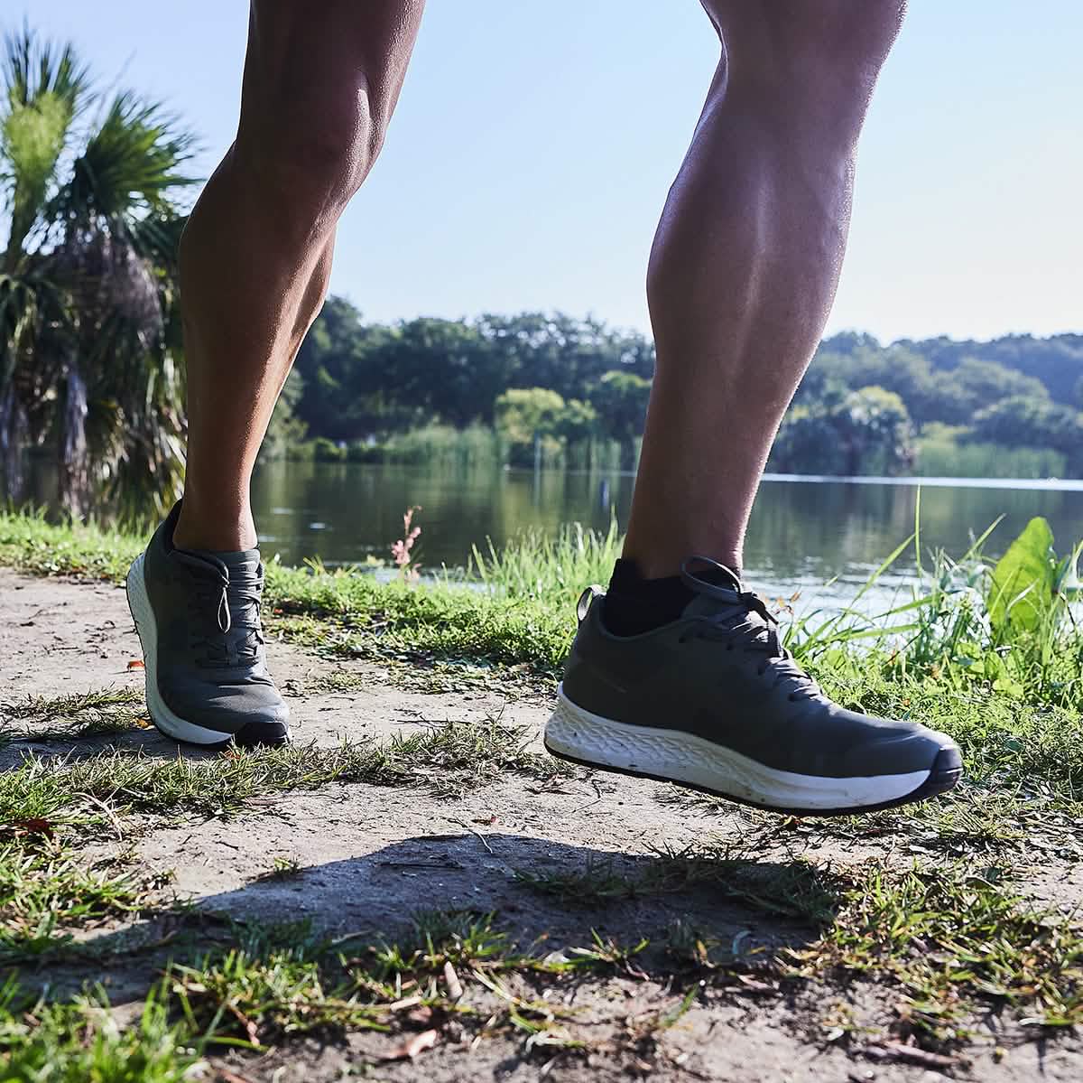 A jogger wearing GORUCK's Men's Rough Runner - Earth shoes runs along a dirt path next to a scenic lake, surrounded by lush greenery and trees. The sun brightly illuminates the scene, creating shadows on the ground, while the Gradient Density midsole offers optimal support for each step.