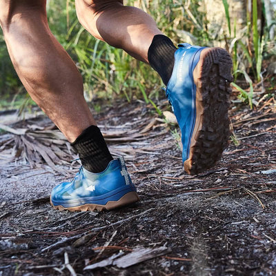 A person is running on a dirt path in a forest, wearing GORUCK Mackall - Legion Blue + Gum trail shoes similar to those used in Special Forces training, paired with black socks. The ground is dusty with scattered leaves, while tall grass and trees line the path, suggesting a rugged outdoor setting.