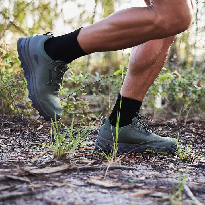 Close-up of a person running on a dirt trail surrounded by greenery, wearing black socks and the GORUCK Mackall - Earth + Forged Iron trail shoes featuring a triple compound outsole. The ground is covered with small plants and dry leaves.