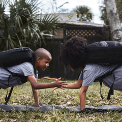 Two kids outdoors doing plank exercises wearing GORUCK backpacks and gray shirts, high-fiving each other on mats