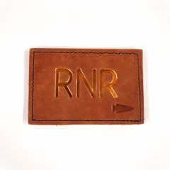 A brown Patch - Custom Leather Monogram featuring "RNR" and a small gold arrow, embossed on leather—ideal as a personalized gift.