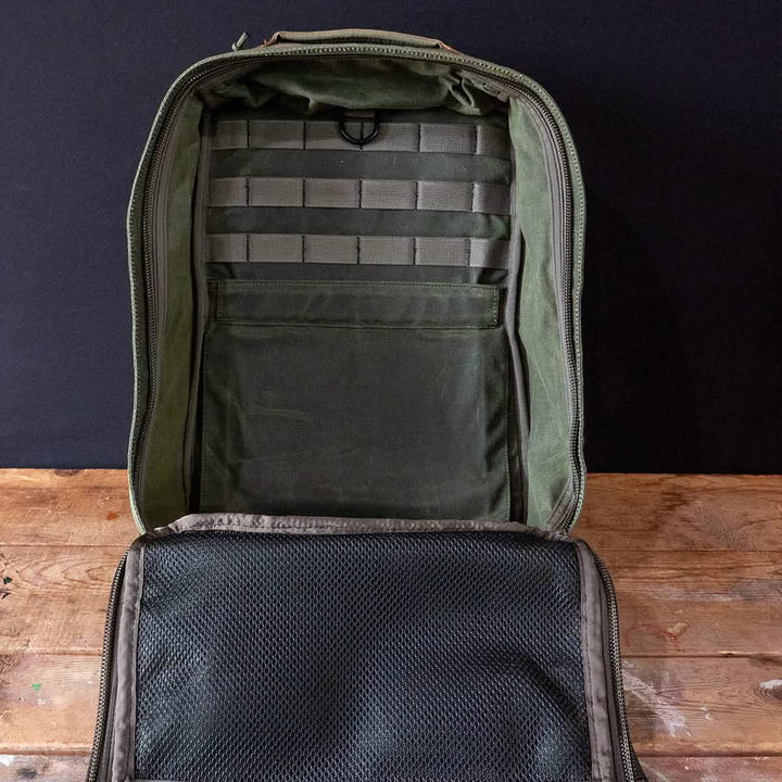 Place the GR1 Heritage - Alligator Leather Front Panel backpack by GORUCK on a wooden surface. The interior features a green hue with various compartments, including mesh pockets and organizational straps. Its retro canvas rucksack design strikingly contrasts against the plain black background.