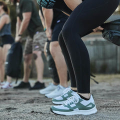 People are outdoors in a line, holding weighted bags and wearing athletic clothing and sneakers, ready for a Rough Runner workout. The focus is on their legs and shoes with Gradient Density EVA Midsoles, suggesting a fitness activity or training session with the Women's Rough Runner - Seaspray by GORUCK.
