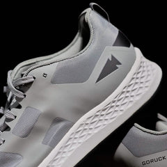 A close-up of the Men's Rough Runner - Speed Grey + White athletic shoe, featuring a gray design with a white textured sole, perfect for the Rough Runner. This GORUCK shoe incorporates mesh fabric and subtle details, along with Gradient Density technology, and displays a triangular logo. The brand name is prominently visible on the heel against a dark background.