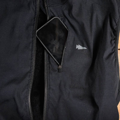 A smartphone partially tucked into a zippered pocket of a sleek black GORUCK Vest of Power - ToughDry® + Fleece, featuring a small white logo on the chest.