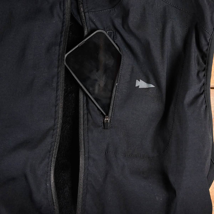 A smartphone partially tucked into a zippered pocket of a sleek black GORUCK Vest of Power - ToughDry® + Fleece, featuring a small white logo on the chest.