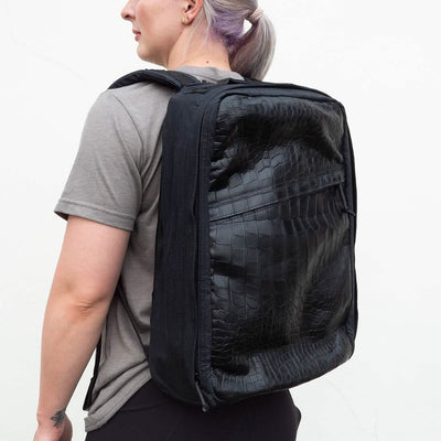 A person with light hair is wearing a gray t-shirt and black pants, carrying a spacious GORUCK GR1 Heritage backpack featuring an alligator leather front panel. The black backpack, worn over both shoulders, complements the retro canvas rucksack style. The background is a plain, light-colored wall.