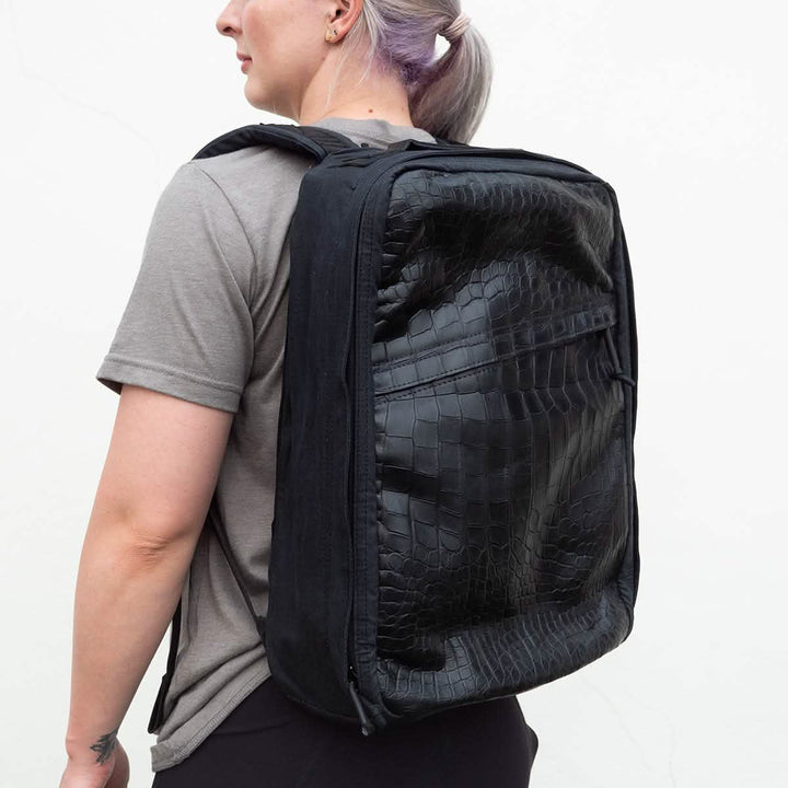A person with light hair is wearing a gray t-shirt and black pants, carrying a spacious GORUCK GR1 Heritage backpack featuring an alligator leather front panel. The black backpack, worn over both shoulders, complements the retro canvas rucksack style. The background is a plain, light-colored wall.