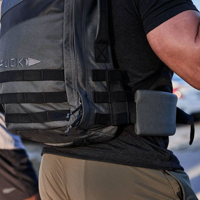Close-up of a person wearing a dark GORUCK backpack featuring MOLLE straps and a rectangular attachment on the side. The Padded Hip Belt by GORUCK ensures comfort for heavy loads. The individual is dressed in a black t-shirt and olive pants, standing outdoors against a blurred background.