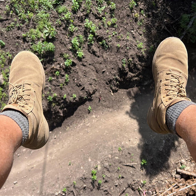 Two feet in tan boots and gray socks dangle above a dirt path with patches of green plants.