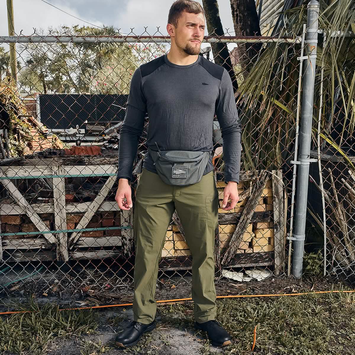 A man stands in front of a chain-link fence and trees, wearing a dark long-sleeve shirt, olive green Tactical FP1 pants by GORUCK, black shoes, and a waist bag with silent zipper pulls. He gazes to the side with a relaxed posture in this outdoor setting with a rustic backdrop.