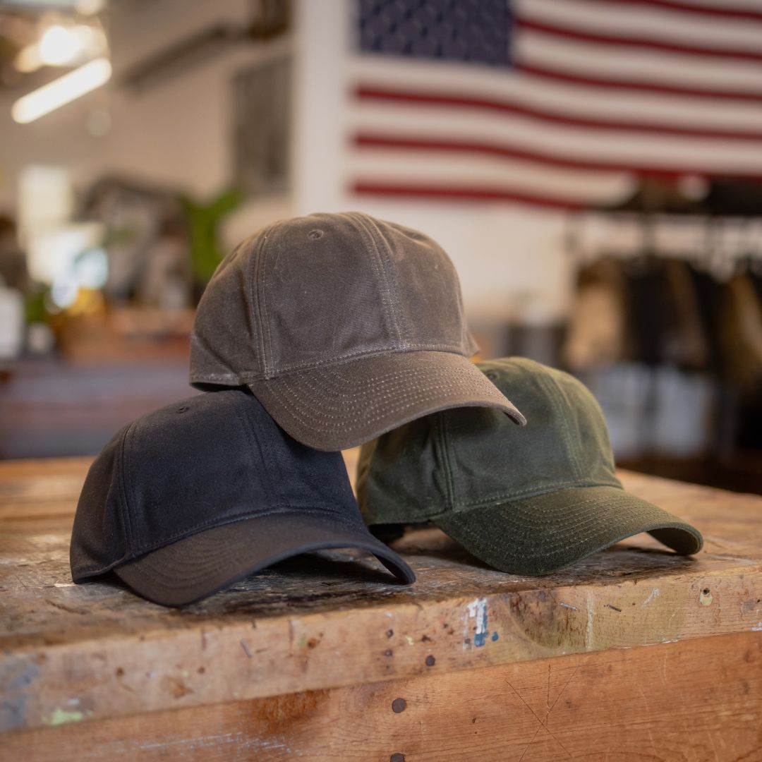 Three Tac Hat - Heritage baseball caps in brown, green, and black are displayed on a wooden table, with an American flag in the background.