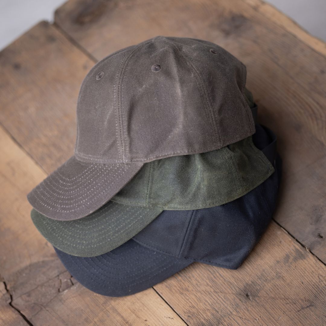 Three Tac Hat - Heritage baseball caps in brown, green, and black are stacked on a rustic wooden surface.