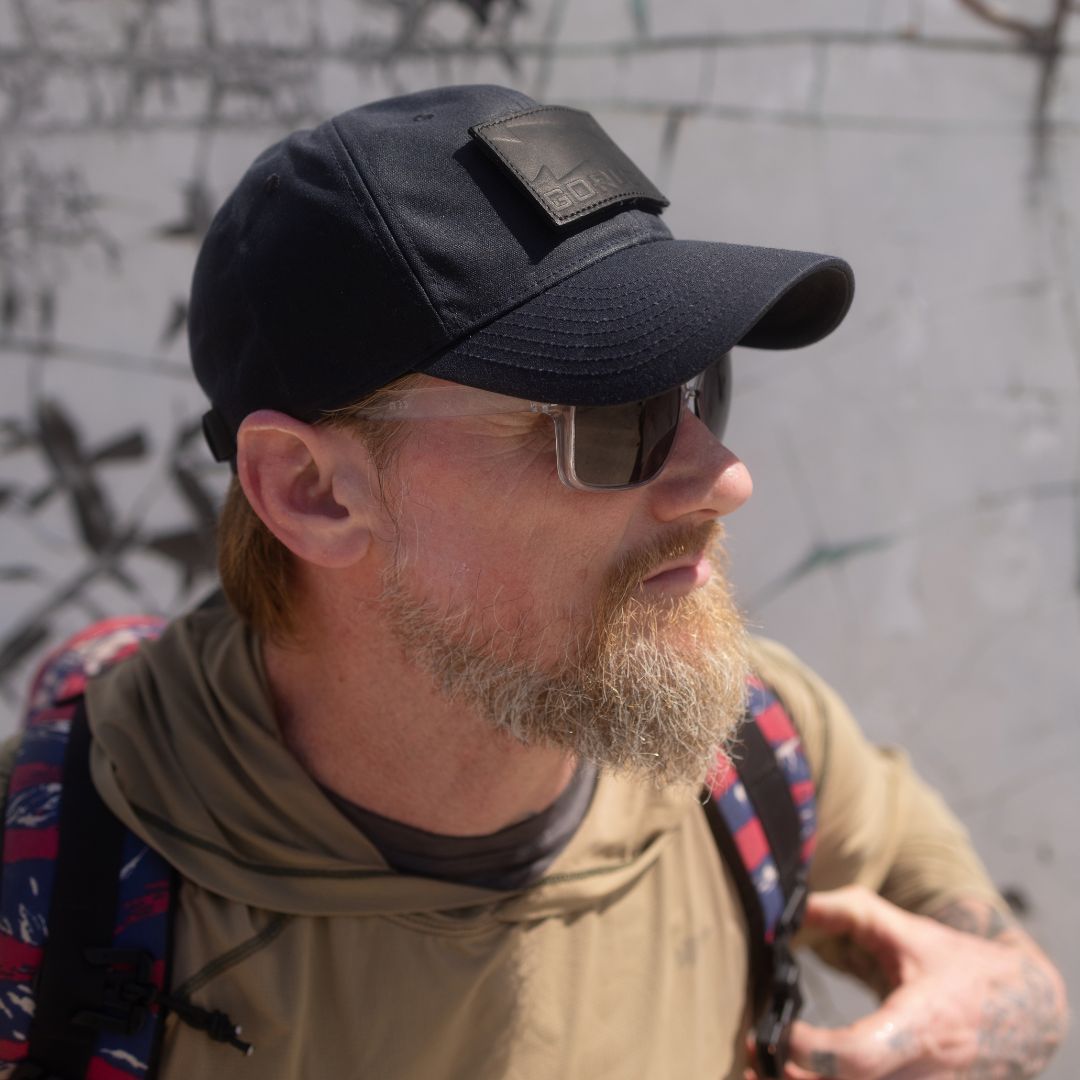 A bearded man wearing sunglasses, the Tac Hat - Heritage, and a backpack stands outdoors near a graffiti wall.