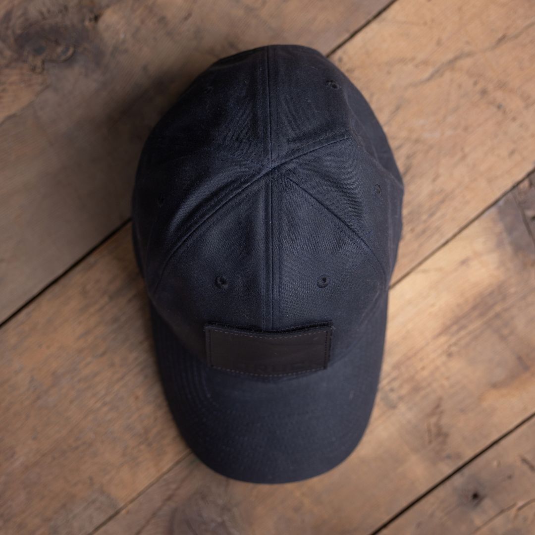 A Tac Hat - Heritage rests on a wooden floor, featuring a black design with a blank patch.
