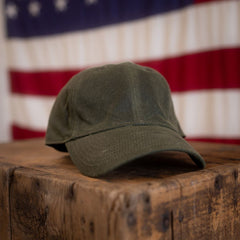 A Tac Hat - Heritage in olive green, displayed on a wooden box with a blurred American flag in the background.