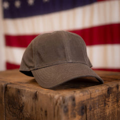 The Tac Hat - Heritage sits atop a rustic wooden box, with a blurred American flag in the background.