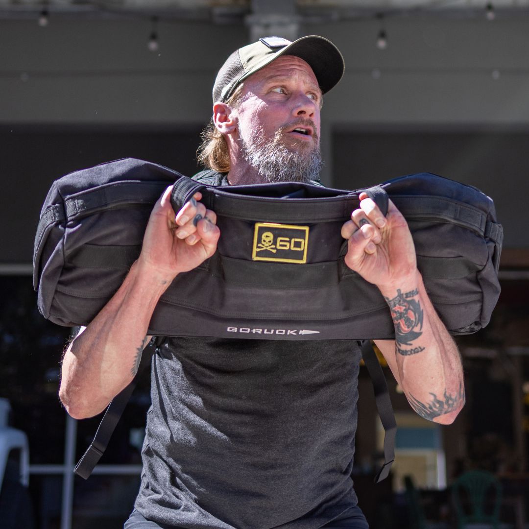 A bearded man wearing a GORUCK Tac Hat performs a front-carry exercise with a black 500D CORDURA sandbag featuring a skull-and-crossbones patch.