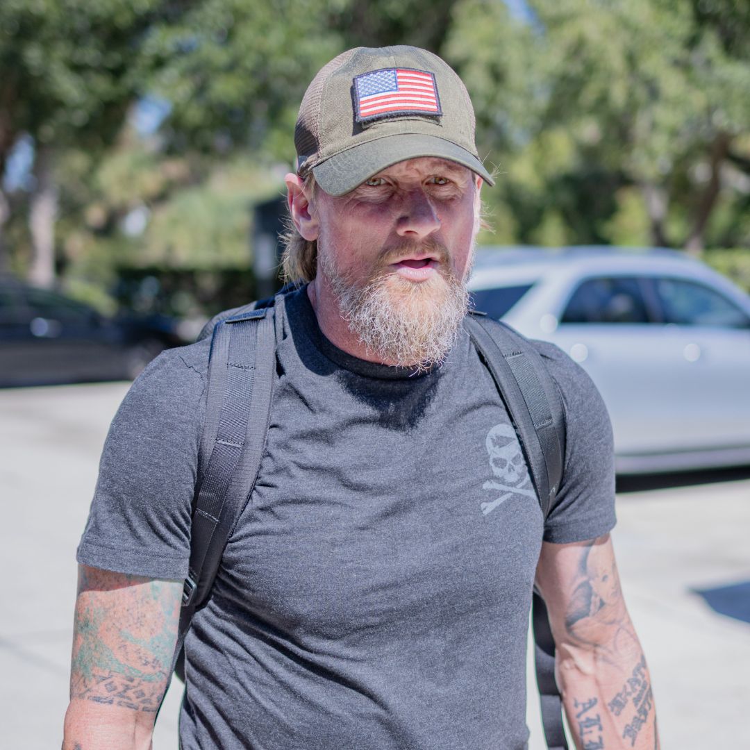 A bearded man with tattoos on his arms wears a dark grey skull t-shirt, a backpack, and an olive green GORUCK Tac Hat with an American flag patch.