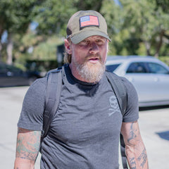 A bearded man with tattoos on his arms wears a dark grey skull t-shirt, a backpack, and an olive green GORUCK Tac Hat with an American flag patch.