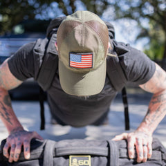 A person with tattooed arms wearing a GORUCK Tac Hat performs a push-up while carrying a backpack.