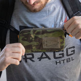 A person holds the Sternum / Belt Pouch - CORDURA in camo on their chest while wearing a gray shirt with “FRAGG HTBL” text, keeping their essentials hands-free—ideal for rucking or outdoor adventures.