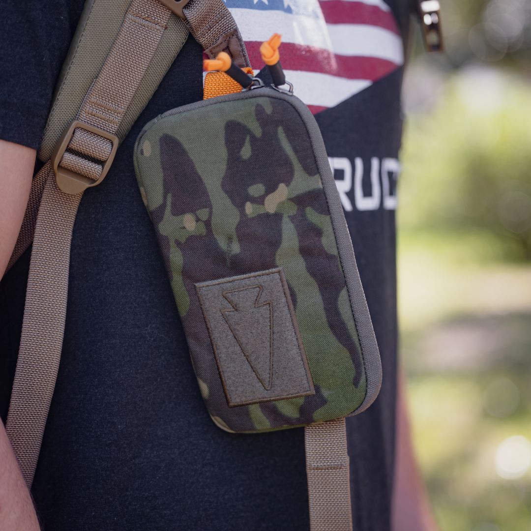 A Sternum / Belt Pouch - CORDURA in camouflage with a patch is attached to a backpack strap worn by a person outdoors, offering a convenient, hands-free solution perfect for rucking adventures.