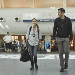 Two people stroll through the airport terminal, signs above pointing to All Gates and Concourses A, B, C. The woman, in a light shirt and jeans, expertly maneuvers her carry-on compliant bag while the man in a dark shirt manages a GORUCK GR2 backpack with Special Forces quality precision. Other travelers bustle around them.