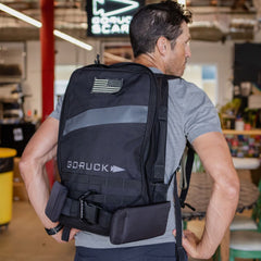 A man wears a black GORUCK MOLLE rucksack with a reflective stripe, an American flag morale patch, and a heavy-duty carabiner attached to the bottom.