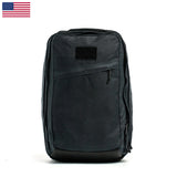 The GR2 - Heritage Waxed Canvas backpack in black features a front zipper pocket and a US flag at the top left, inspired by rugged travel rucks, displayed on a white background.