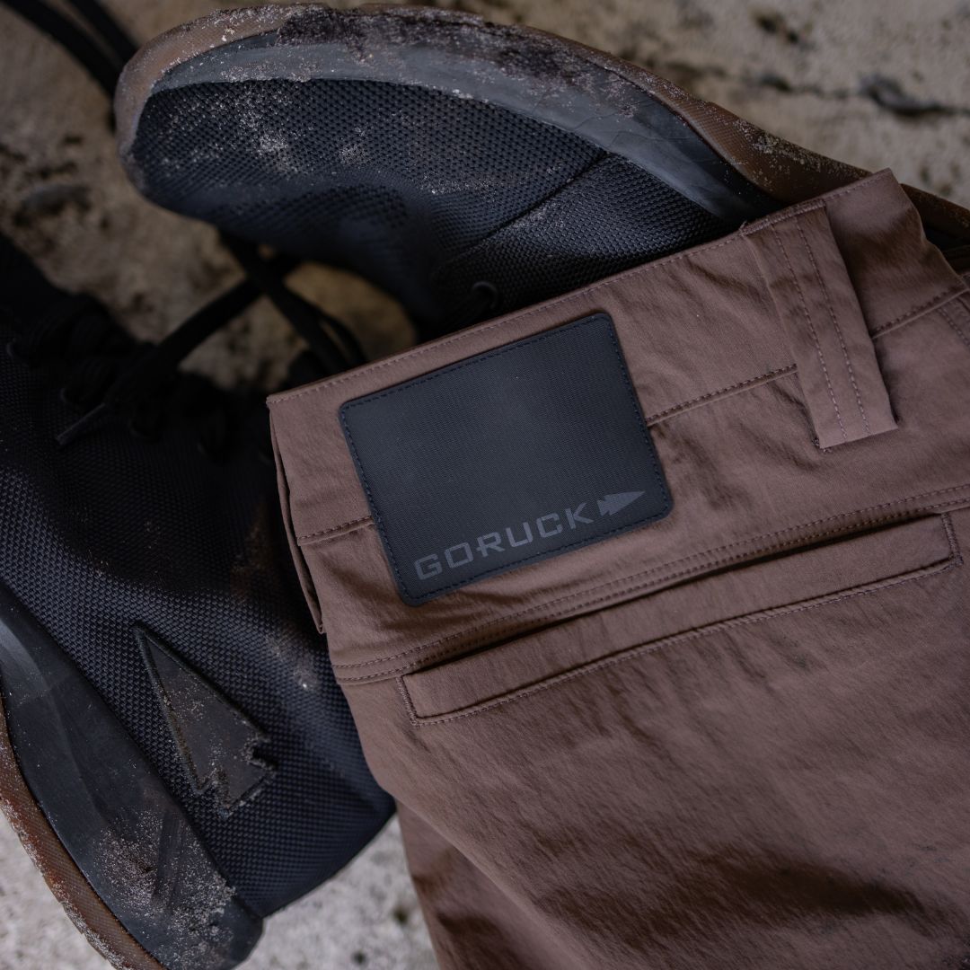 Brown GORUCK abrasion resistant pants with a black brand patch, resting next to a pair of black athletic shoes.