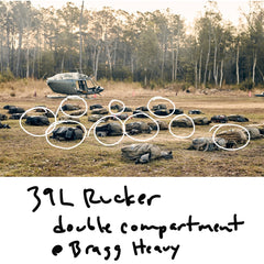 Many endurance rucksacks are scattered across a field in front of a parked helicopter, with several circled in white. Handwritten text below reads “39L Rucker double compartment Bragg Heavy.”