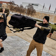 Two men training outdoors with the Rucker - Long Range, lifting heavy bags overhead on a cloudy day—ideal for distance rucking or prepping for upcoming rucking events.