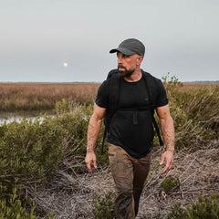A man in outdoor clothing and a cap, carrying the Rucker - Long Range rucksack, walks through marshy grassland at dusk with a serious expression.