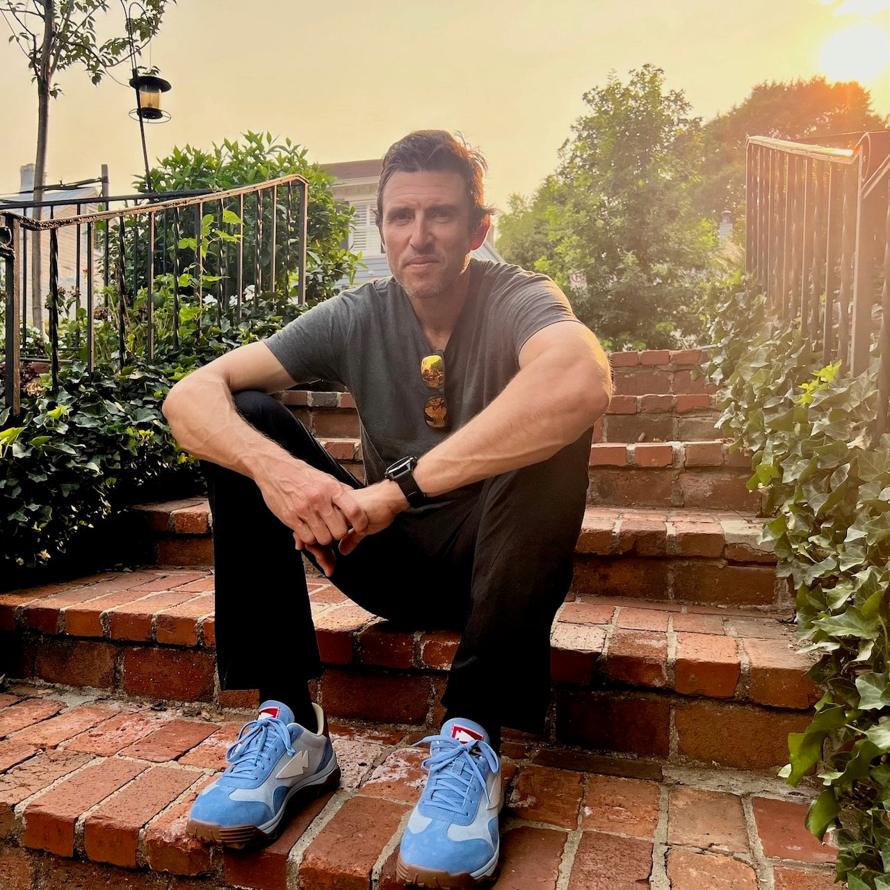 A man sits on brick steps outdoors at sunset, wearing a gray shirt, black pants, and the Mackall 45 Americana blue sneakers.