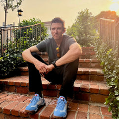 A man sits on brick steps outdoors at sunset, wearing a gray shirt, black pants, and the Mackall 45 Americana blue sneakers.
