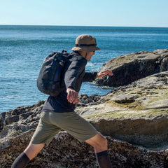 Carrying the Mesh Ruck, someone treks across rocky coastal terrain beneath a clear blue sky.