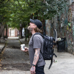 Man with beard wearing GORUCK backpack carrying skateboard and coffee in urban alleyway