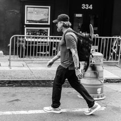 Man walking with GORUCK backpack carrying skateboard, urban street scene, durable rucking gear