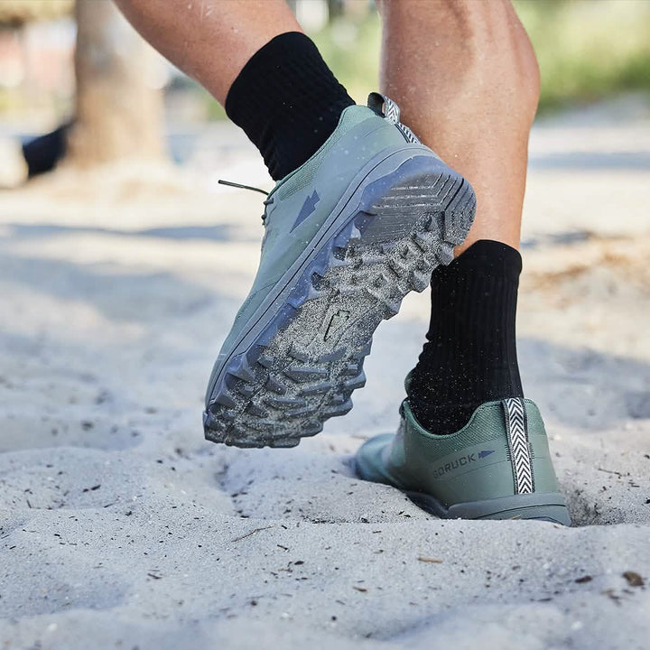 A person running on a sandy surface wears GORUCK's Mackall trail shoes in Earth and Forged Iron colors with black socks. The shoes feature a triple compound outsole for unmatched grip, set against a backdrop of blurred trees and a bright sky.