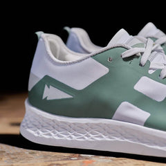 Close-up of the Men's Rough Runner - Seaspray sneaker by GORUCK, showcasing a green and white design. The shoe displays geometric patterns and a stylized lightning bolt graphic on the side. It boasts a textured white sole crafted from Gradient Density EVA, complemented by light gray laces, set against a dark background.