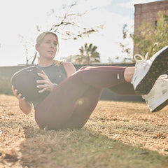 Exercising outdoors on the grass, a person holds a Sand Medicine Ball by GORUCK, twisting to enhance core stability. Under the bright sunlight casting a warm glow, they sport a sleeveless top, maroon leggings, and white sneakers.