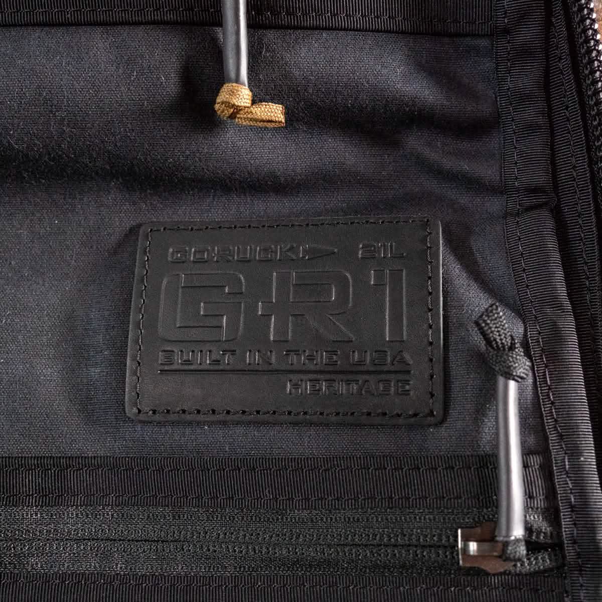 Close-up of a black alligator leather patch on a retro canvas rucksack, embossed with the words "GORUCK GR1 Heritage - Built in the USA" alongside the volume measurement of 21L. The patch is surrounded by black fabric and zippers.