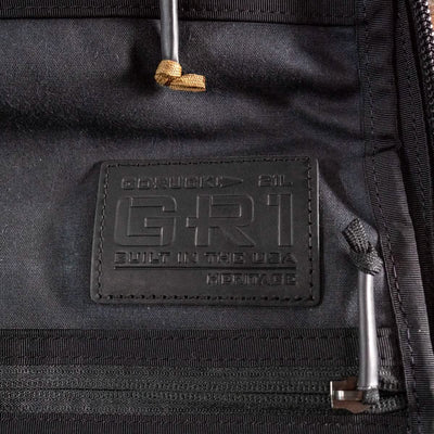 Close-up of a black alligator leather patch on a retro canvas rucksack, embossed with the words "GORUCK GR1 Heritage - Built in the USA" alongside the volume measurement of 21L. The patch is surrounded by black fabric and zippers.