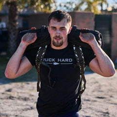 Man carrying black rucking sandbag on shoulders outdoors, wearing black "RUCK HARD" t-shirt, tactical backpack