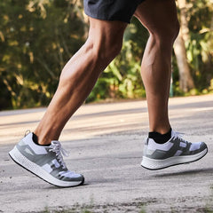 A man wearing the Men's Rough Runner shoes in White and Speed Grey from GORUCK, featuring a Gradient Density™ EVA Midsole, strolls along a sunlit path surrounded by greenery. Dressed in dark shorts, his muscular legs stand out as he enjoys the serenity of outdoor exercise.