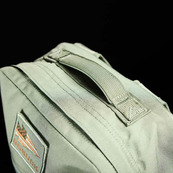 Close-up of the KR1 kids rucksack by GORUCK in green fabric, complete with a woven handle and padded straps. It includes a patch featuring triangular designs and the text "GORUCK" in orange. The black background highlights the bag's details, making it perfect for carrying school supplies.