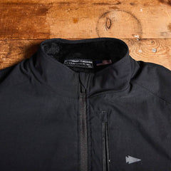 A close-up of the GORUCK Vest of Power - ToughDry® + Fleece in black, with a zippered front, is displayed on a wooden surface. The high-collar design and interior label featuring an American flag tag highlight its sturdy craftsmanship, complemented by a subtle arrow logo that signifies the Vest of Power.