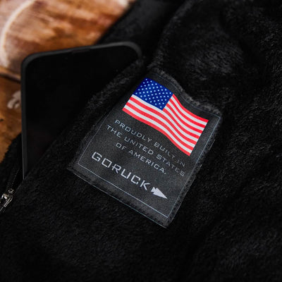 Close-up of a label on black fabric displaying the American flag and text Proudly Built in the United States of America above the GORUCK brand name. Next to the label, a smartphone is partially visible on a wooden surface, featuring GORUCK's Vest of Power - ToughDry® + Fleece commitment.